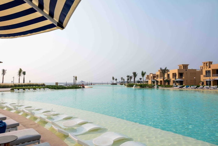 Spacious infinity pool with in-water loungers and waterfront villas in the background under a clear sky