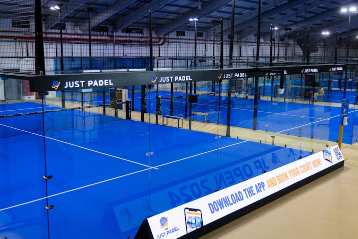 Indoor courts showcasing bright blue playing surfaces, glass walls, and a banner encouraging players to download the app to book courts, all within a modern, well-lit sports facility.