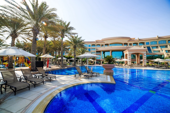 Spacious outdoor pool area at a resort with palm trees, sun loungers, and a large hotel building in the background under clear skies.