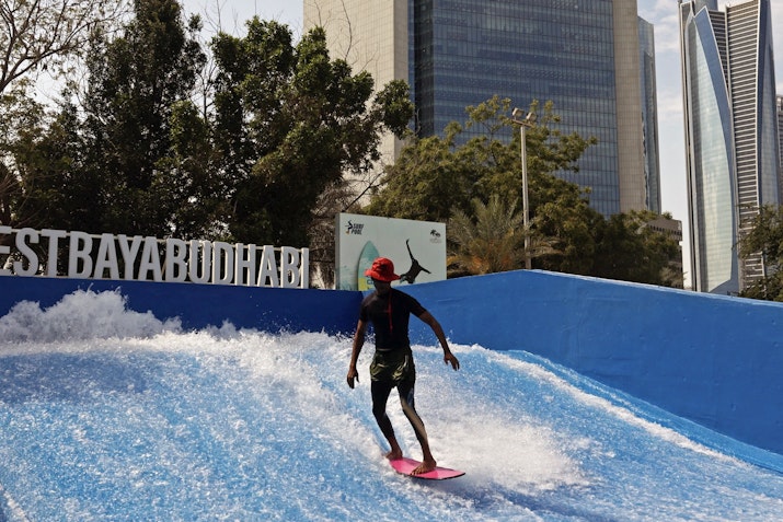 Person surfing on an artificial wave with city buildings in the background.