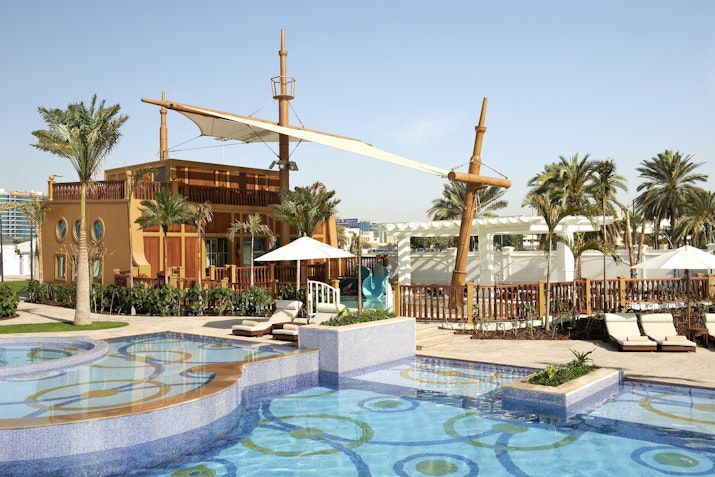 Resort pool with circular blue patterns, palm trees, lounge chairs, and a pirate ship structure in the background.
