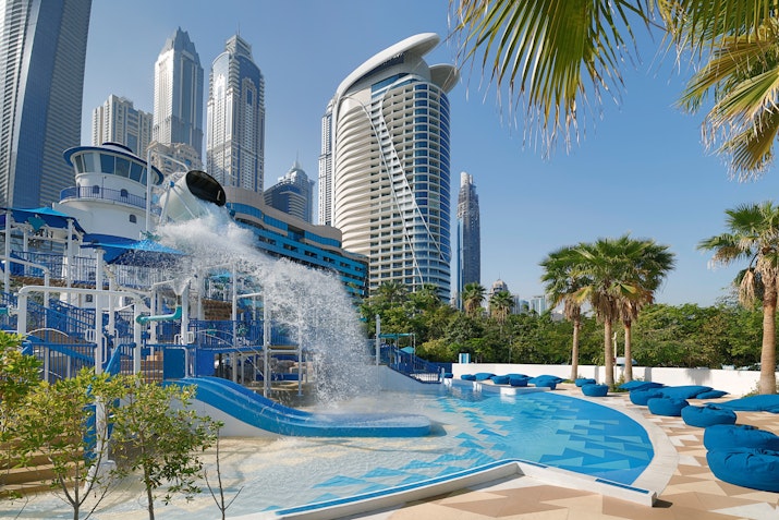 Modern outdoor pool area with blue loungers, palm trees, and a city skyline in the background, featuring a unique water park attraction.