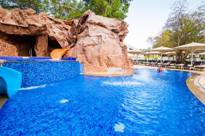 Pool with a rock feature and water slide, surrounded by lounge chairs and umbrellas.