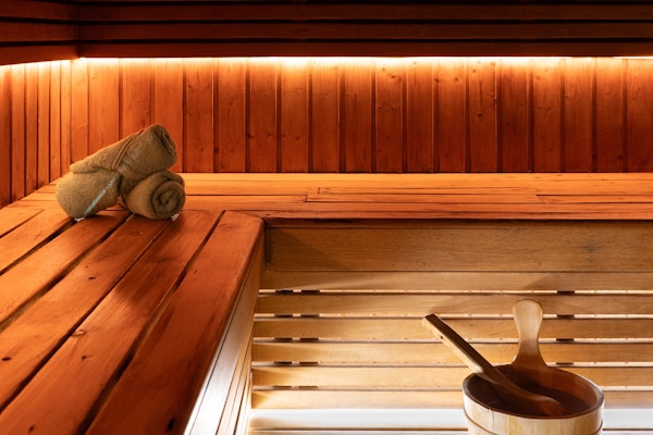 Wooden sauna interior with warm ambient lighting, rolled towels, and a wooden water ladle and bucket.