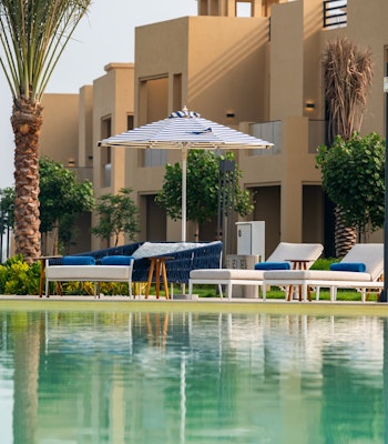 Sofitel Al Hamra Beach Resort A serene view of a resort with a reflective pool in the foreground, lounge chairs, and palm trees, featuring beige buildings in the background.