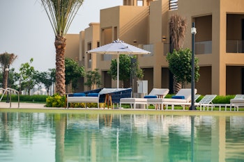 A serene view of a resort with a reflective pool in the foreground, lounge chairs, and palm trees, featuring beige buildings in the background.