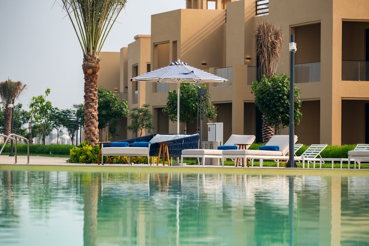 A serene view of a resort with a reflective pool in the foreground, lounge chairs, and palm trees, featuring beige buildings in the background.