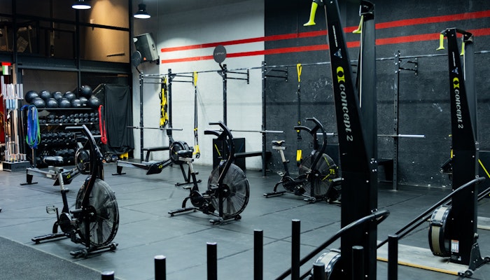 The Project DXB A gym area featuring Concept2 machines, air bikes, TRX suspension trainers, and a variety of fitness equipment against a black and red accent wall, with neatly organized medicine balls and resistance bands in the background.