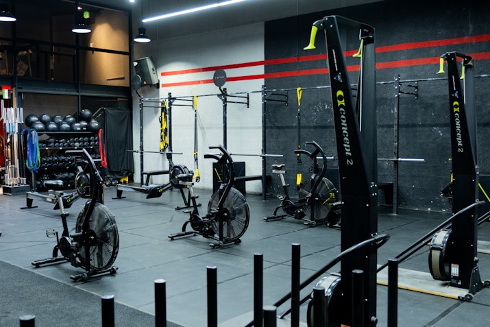 A gym area featuring Concept2 machines, air bikes, TRX suspension trainers, and a variety of fitness equipment against a black and red accent wall, with neatly organized medicine balls and resistance bands in the background.