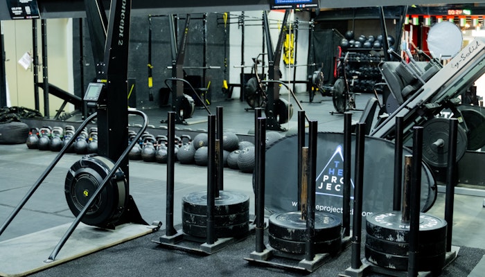 The Project DXB A gym setup featuring sled push weights, Concept2 rowing machines, kettlebells, and various fitness equipment, organized in a workout area with black rubber flooring and equipment racks.