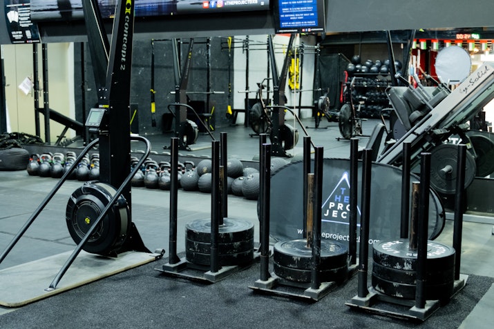 A gym setup featuring sled push weights, Concept2 rowing machines, kettlebells, and various fitness equipment, organized in a workout area with black rubber flooring and equipment racks.