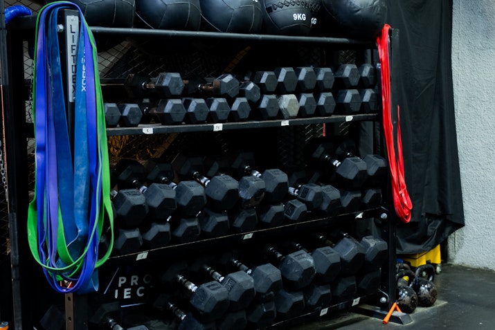 A gym rack stocked with a variety of hex dumbbells, resistance bands in different colors, and medicine balls, organized for easy access in a workout area.