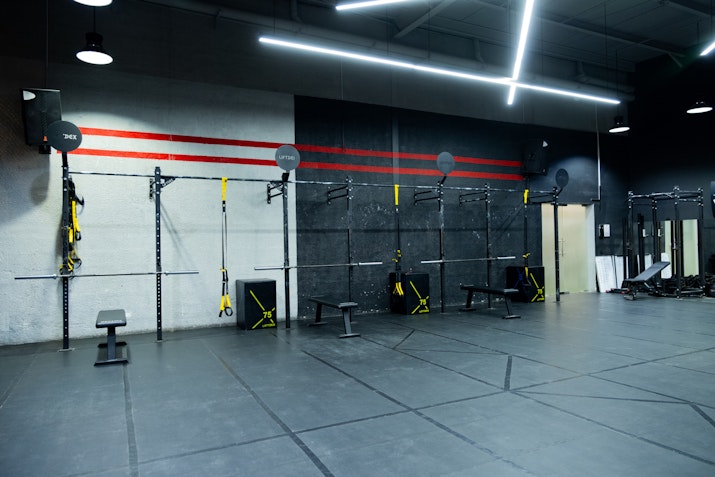 Gym setup featuring TRX suspension trainers, wall balls, benches, and multiple workout stations against a black and gray wall with red accents, designed for functional and strength training