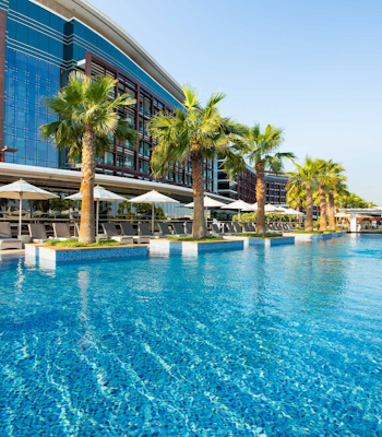 Marriott Hotel Al Forsan (Olympic size lap pool) Outdoor pool with palm trees and a modern building in the background.