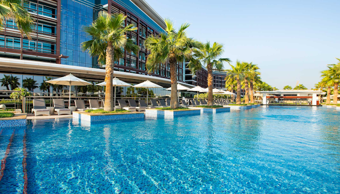Marriott Hotel Al Forsan (Olympic size lap pool) Outdoor pool with palm trees and a modern building in the background.