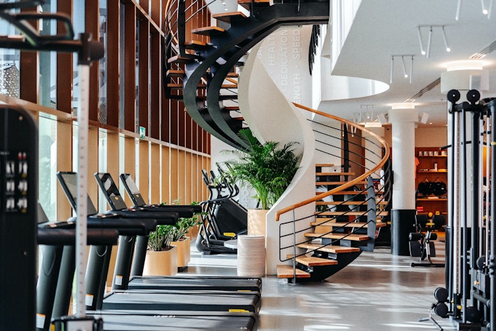 Stylish gym interior featuring treadmills, strength equipment, and a central spiral staircase with surrounding greenery, creating a refined and airy atmosphere.
