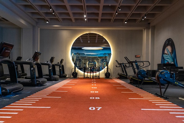 Indoor gym featuring an orange sprint track with markings, surrounded by treadmills and exercise bikes, leading to a circular illuminated mirror on the back wall.