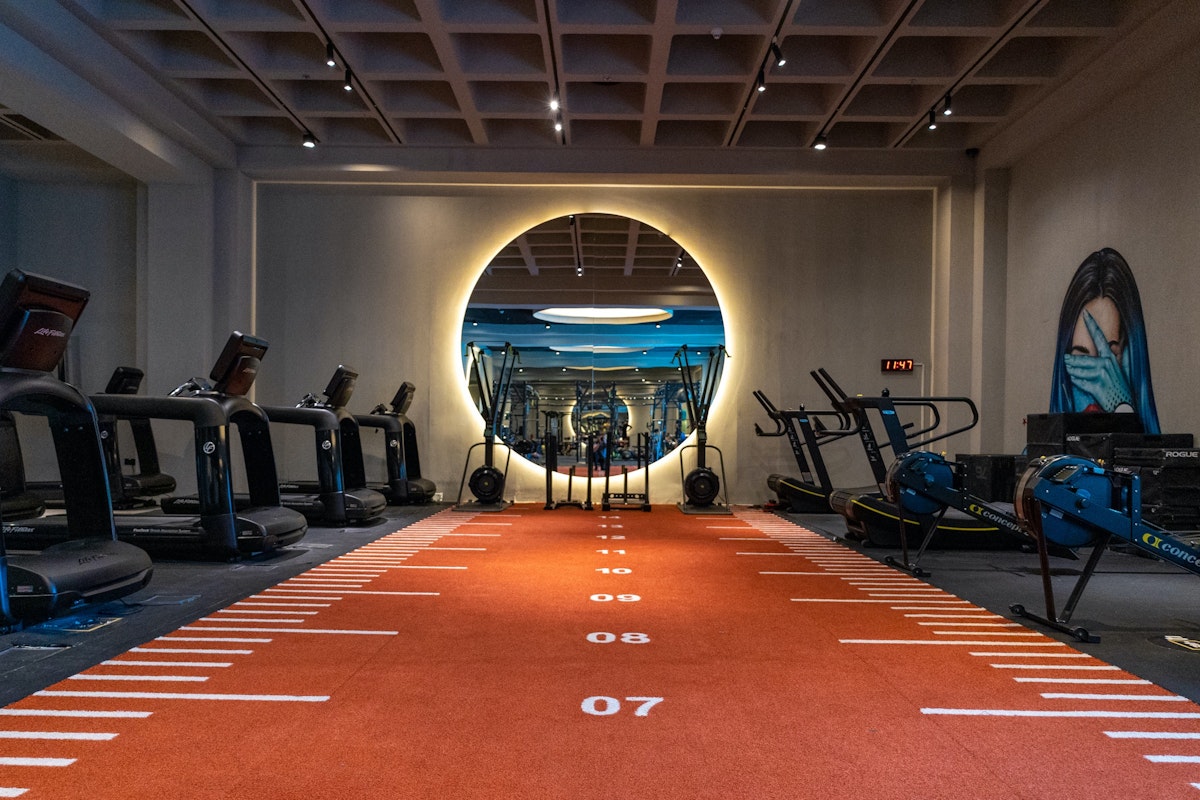 Indoor gym featuring an orange sprint track with markings, surrounded by treadmills and exercise bikes, leading to a circular illuminated mirror on the back wall.