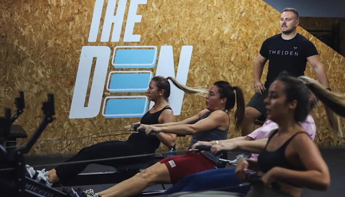 The Den DXB Three women on rowing machines doing a workout with an instructor looking on