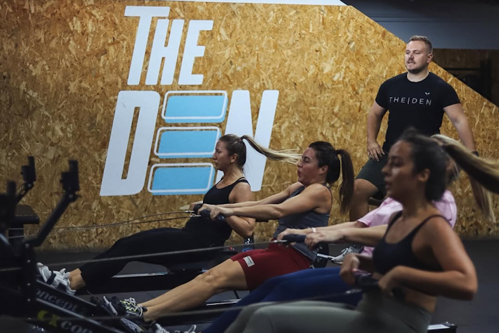 Three women on rowing machines doing a workout with an instructor looking on