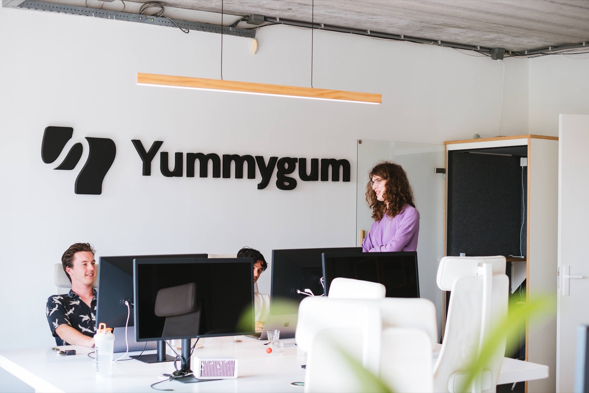 Light office space with desks, chairs, monitors and 3 people talking