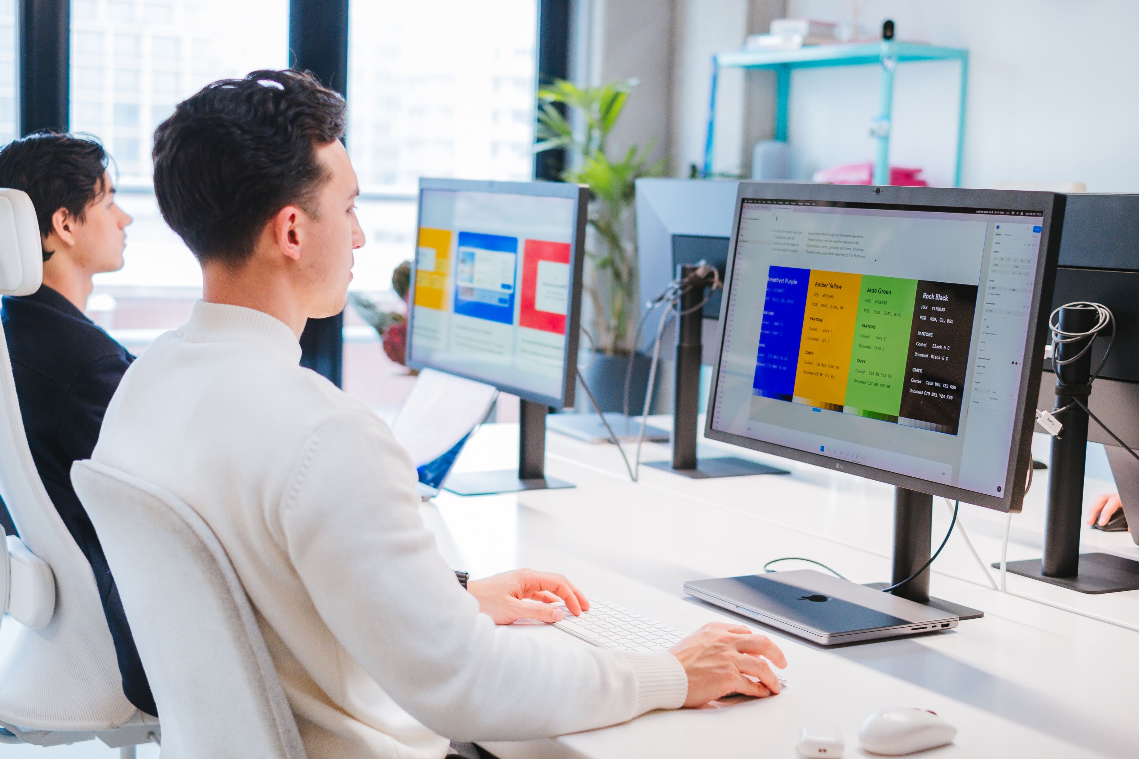 Two men working at a white desk setup in a modern office, each focused on large monitors displaying colorful UI design mockups. The foreground screen shows color-coded sections labeled Amethyst Purple, Amber Yellow, Jade Green, and Rock Black, suggesting a design system or style guide. Bright natural light fills the room, with potted plants and shelves in the background.
