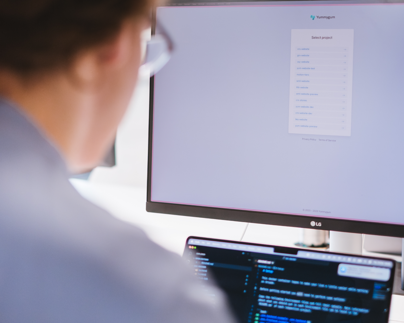 A person is sitting at a desk, working on a computer with two monitors. The larger monitor displays a project selection screen, and the smaller one shows lines of code. The person is wearing glasses and focusing on the screens.