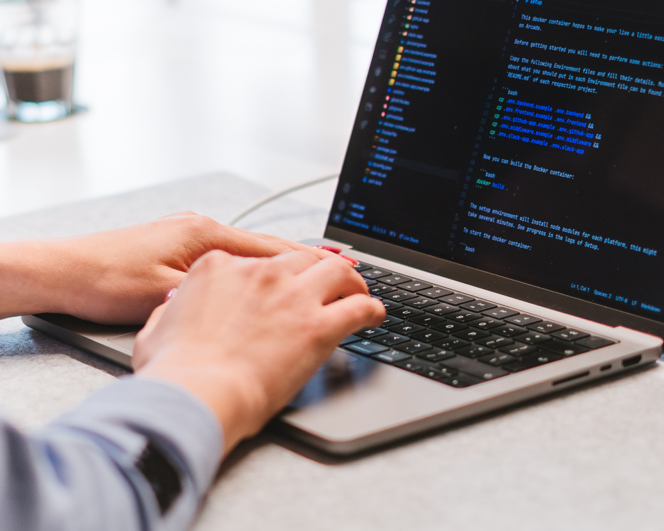 A person typing on a laptop with code displayed on the screen. The workspace is organized, with a notepad and a pen nearby. A blurred coffee mug is in the background. The environment appears calm and focused.
