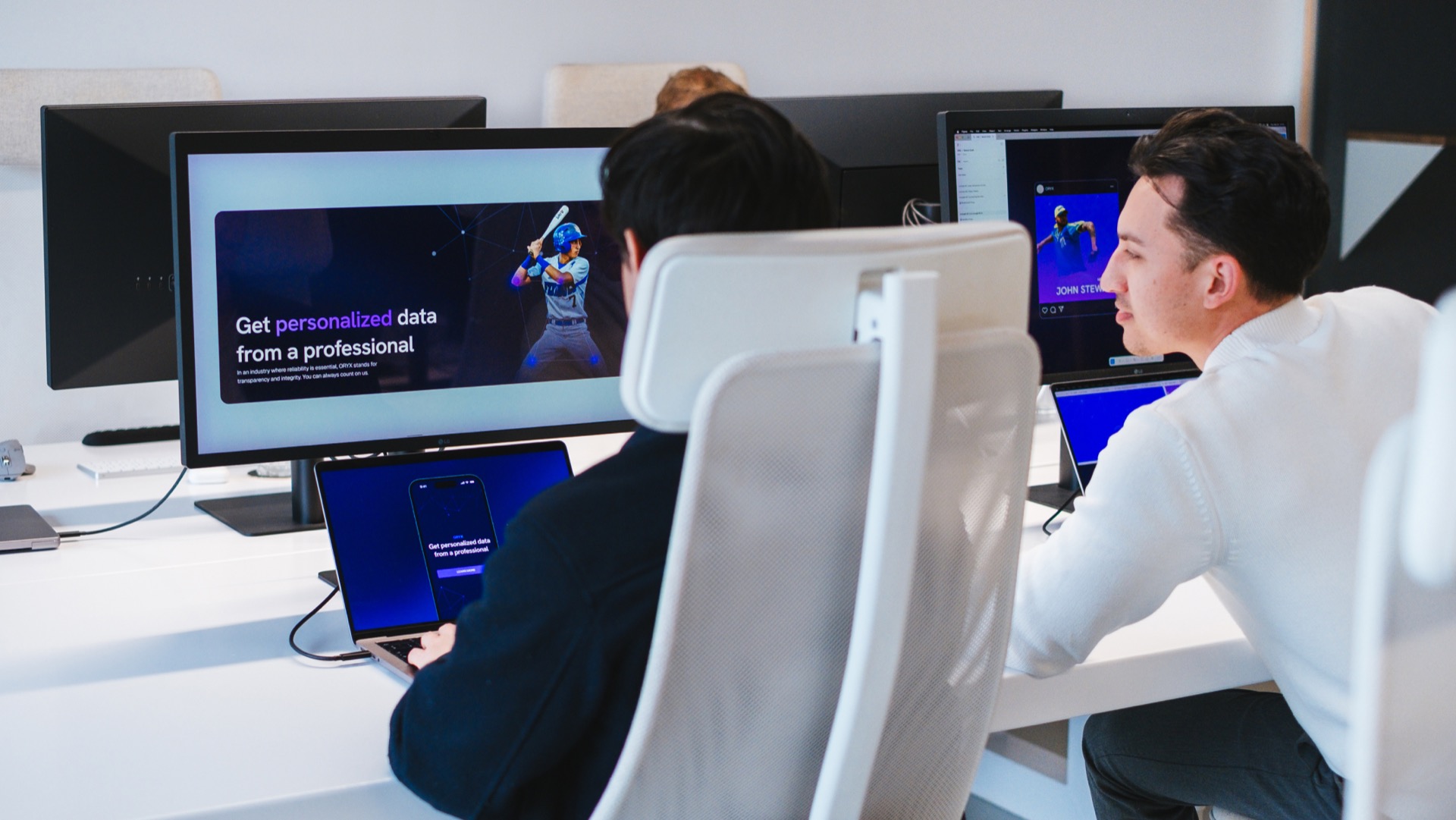 Two people seated at a desk work on laptops and monitor displays. The screens show a digital presentation on personalized data services, featuring a graphic of a person using a device. The office has a modern and clean design.