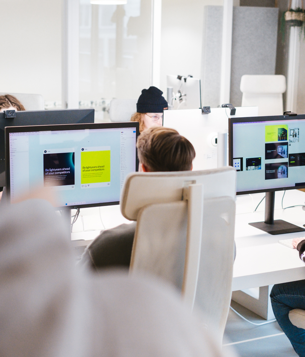 People working at desks with computer monitors in a modern office setting. Monitors display colorful content and social media. One person wears a black beanie. Bright, natural lighting fills the space.