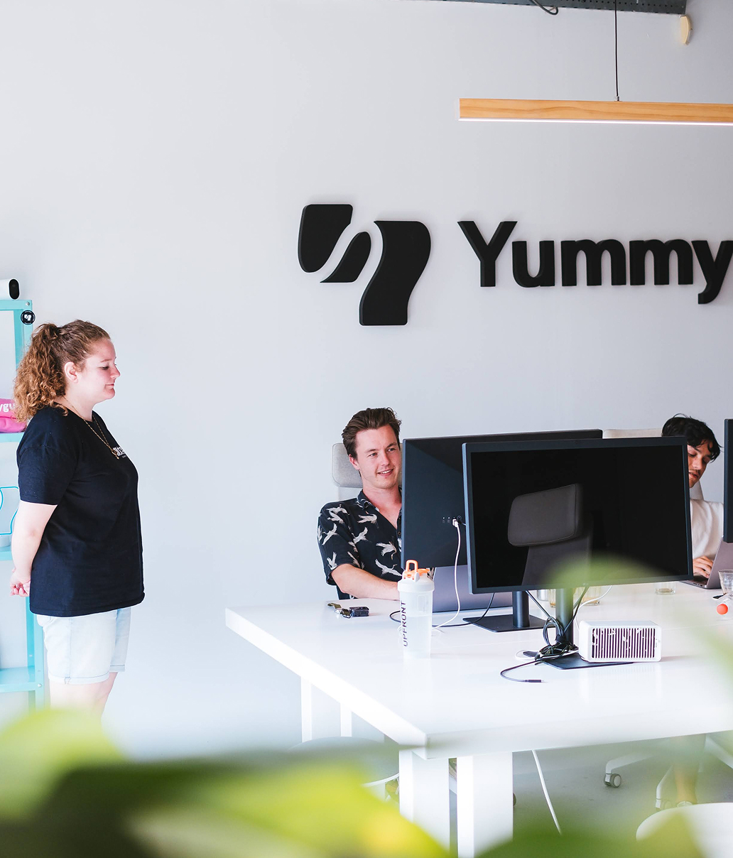 Three people are in a modern office with a "Yummy" logo on the wall. Two men are seated at computers, while a woman stands nearby. The setting is bright with minimalist decor.