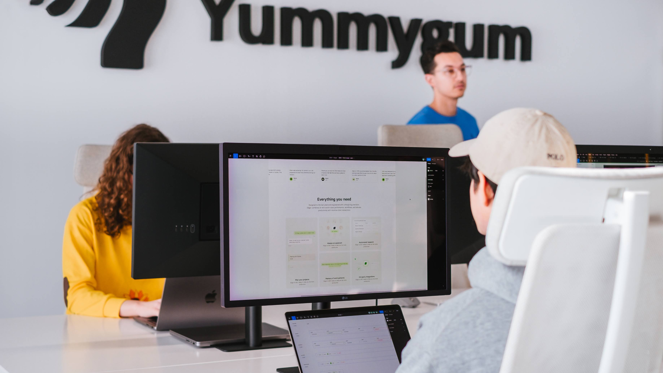 People working at computer desks in an office with a sign reading "Yummygum" on the wall. A large monitor displays a webpage. The workers are focused on their screens, and the atmosphere is casual and collaborative.