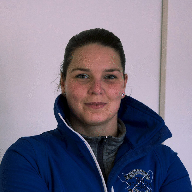 A person wearing a blue jacket with a white logo stands against a plain background. They have short hair and a subtle smile, with their arms crossed.
