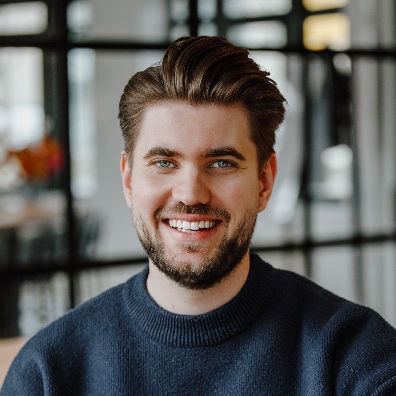 A person with a beard and styled hair smiling, wearing a dark sweater. The background features a modern, blurred indoor setting with black-framed windows.