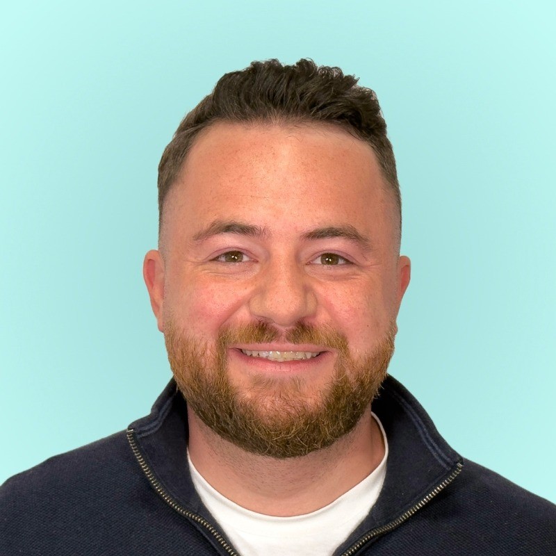 A man with short hair and a beard smiles at the camera. He is wearing a dark zip-up sweater over a white shirt. The background is a solid light teal color.