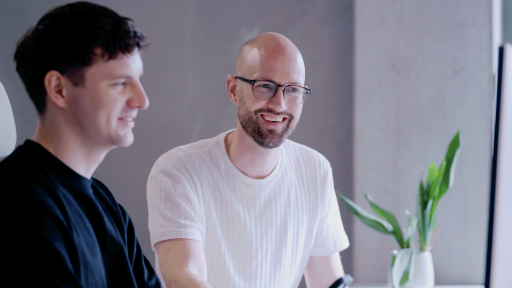 Two people are sitting side by side, engaged in conversation and smiling. One is wearing a black shirt and the other a white shirt and glasses. A green plant is visible in the background against a gray wall.