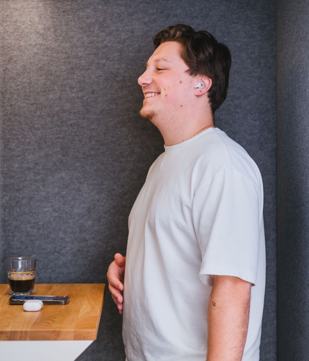 Smiling person in a white shirt leans against a gray wall wearing earbuds. They stand next to a wooden ledge holding a glass of iced coffee, a smartphone, and an open earbud case.