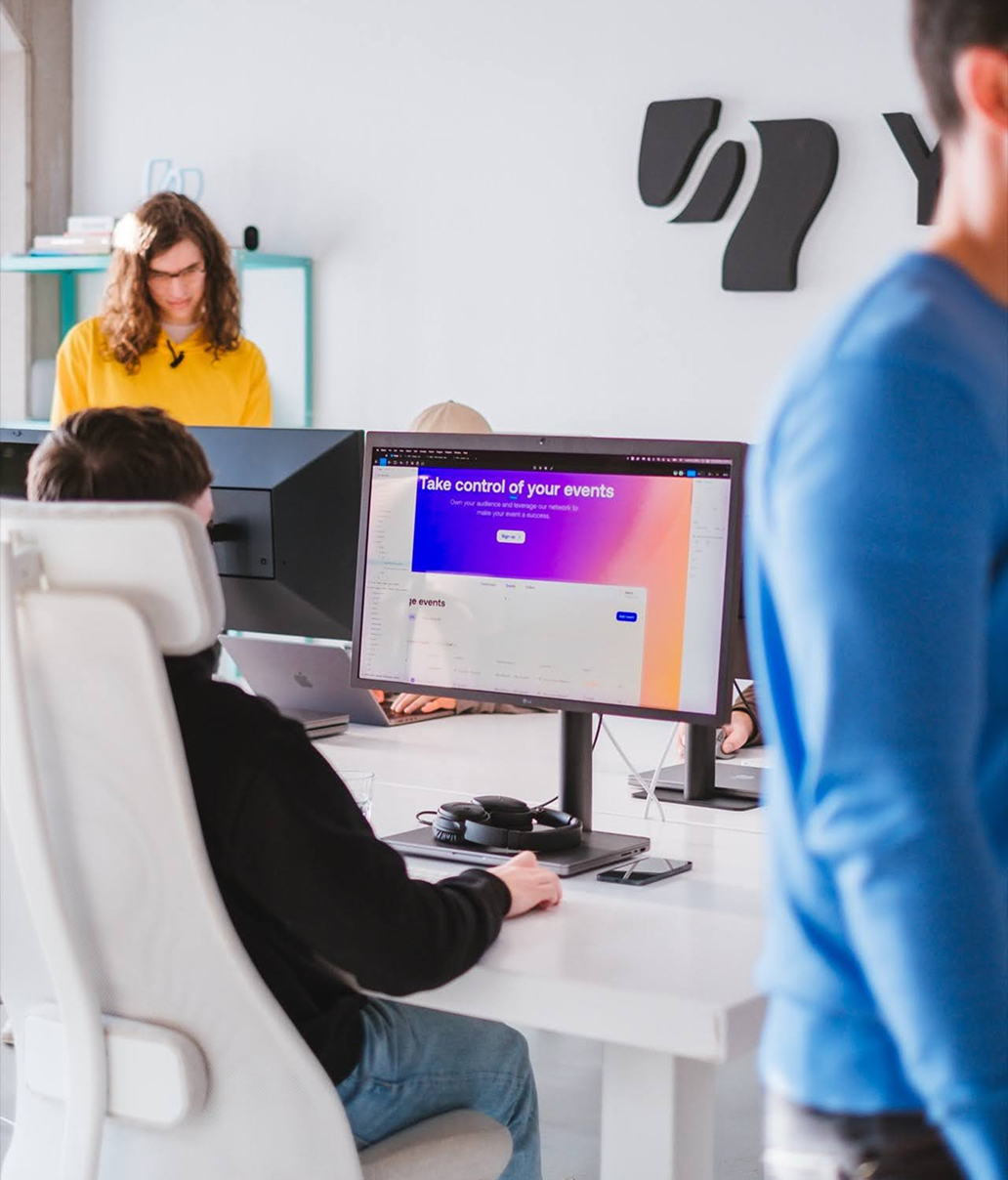 A person sits at a desk working on a computer with a colorful event management website on the screen. Another person with long hair and a yellow shirt stands in the background. A person in a blue shirt walks by. The office is bright and modern.