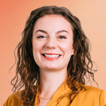 A smiling woman with wavy brown hair, wearing a mustard yellow blazer, stands in front of a warm orange gradient background.