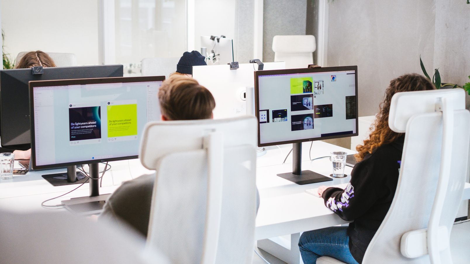 Two people sit at white desks working on desktop computers in a modern, bright office space. Their screens show graphic design projects. The workspace features white chairs, glass partitions, and minimal decor.