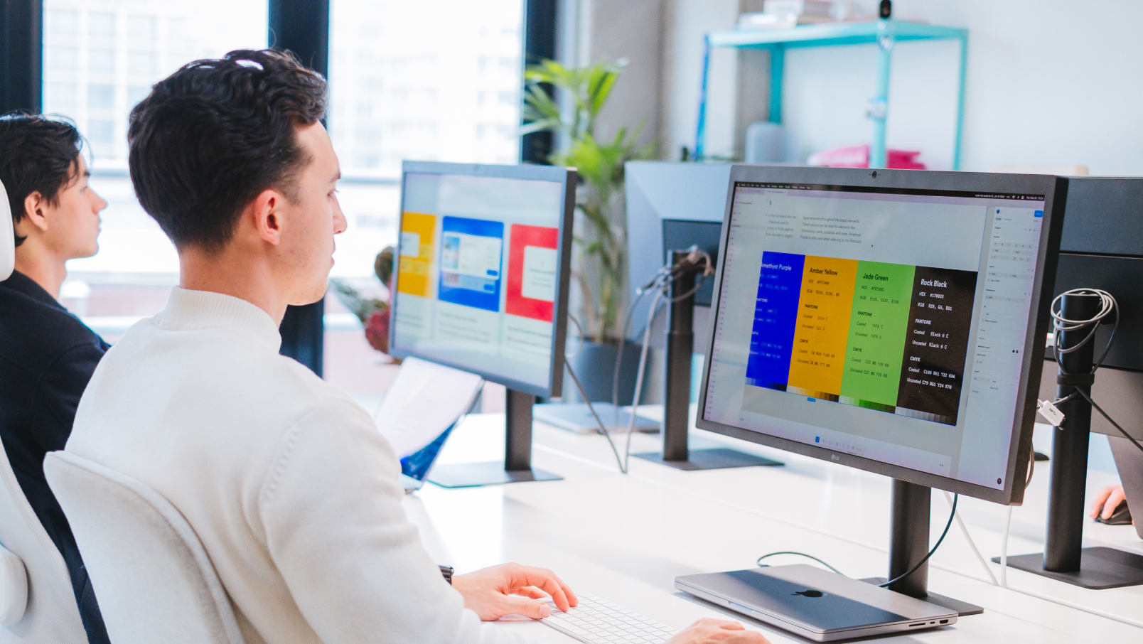 Two people work at desks with large computer monitors displaying colorful charts and documents in a bright, modern office with large windows and plants in the background.