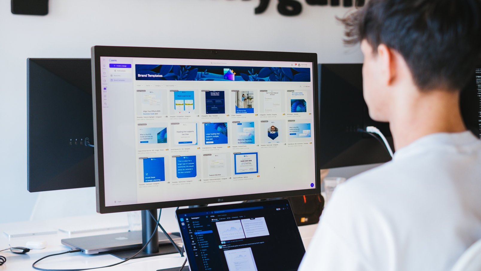 A person sits at a desk using a computer with a large monitor displaying a digital design interface featuring templates and graphics, while a laptop shows code or file directories.