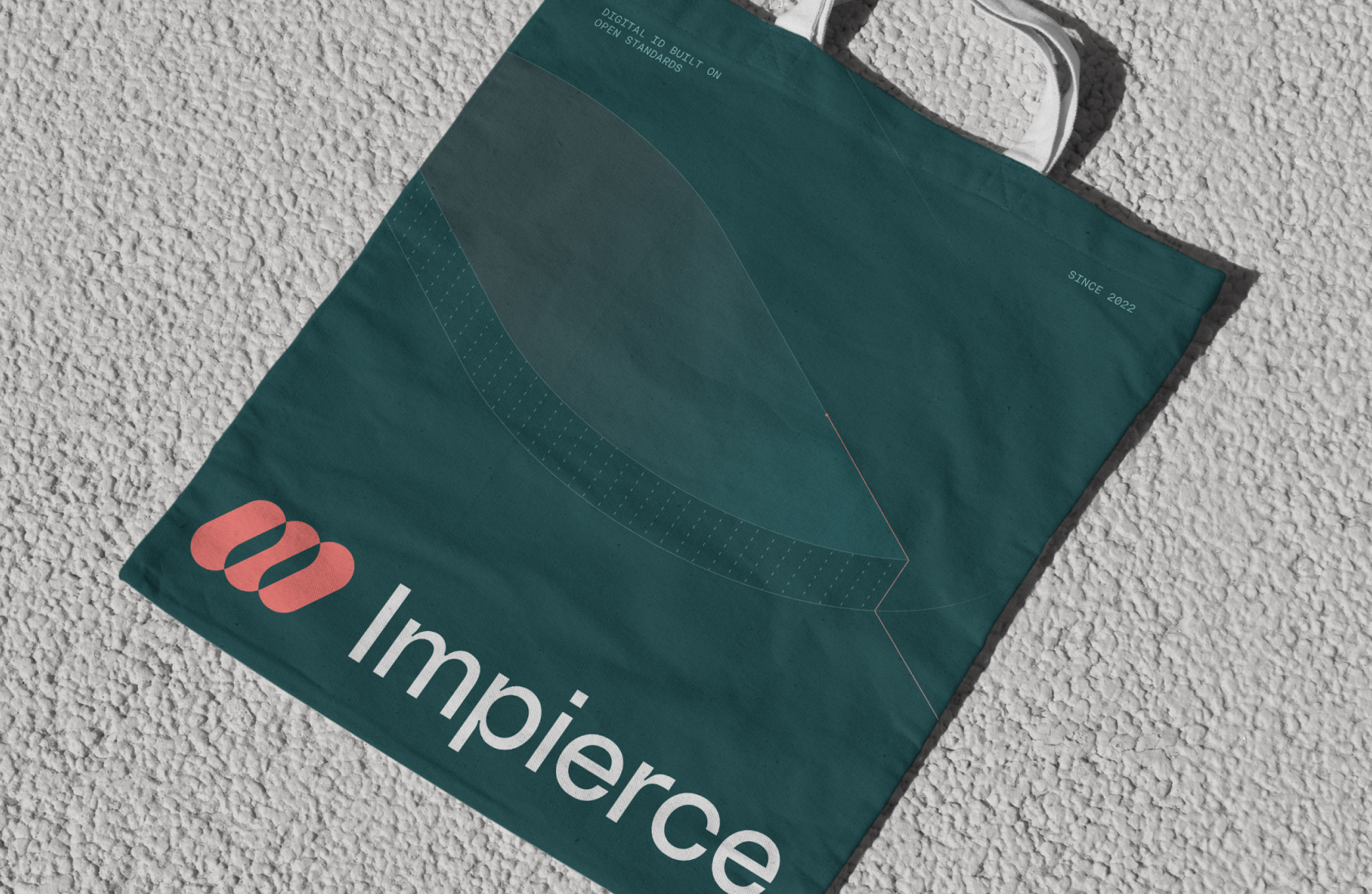 A dark teal tote bag with white handles lies on a textured light surface. The bag features a pink logo and the word "Impierce" printed in bold white letters near the bottom.