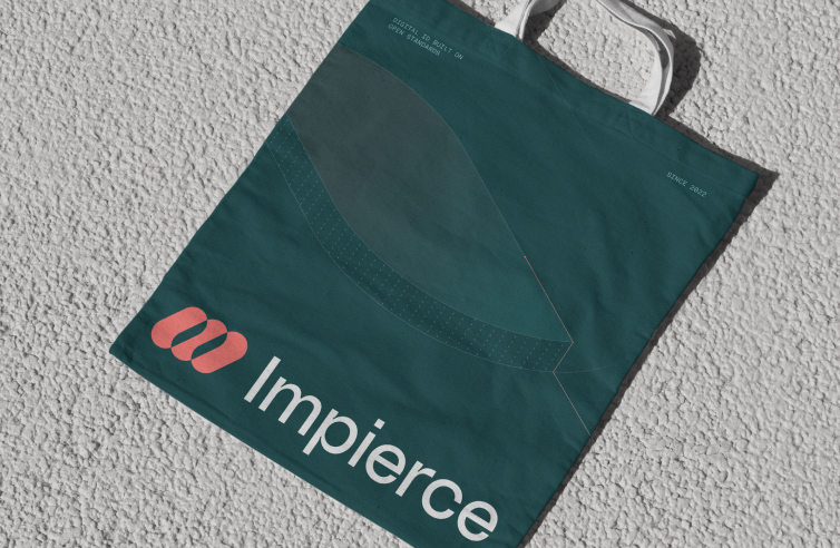 A dark teal tote bag with white handles lies on a textured light surface. The bag features a pink logo and the word "Impierce" printed in bold white letters near the bottom.