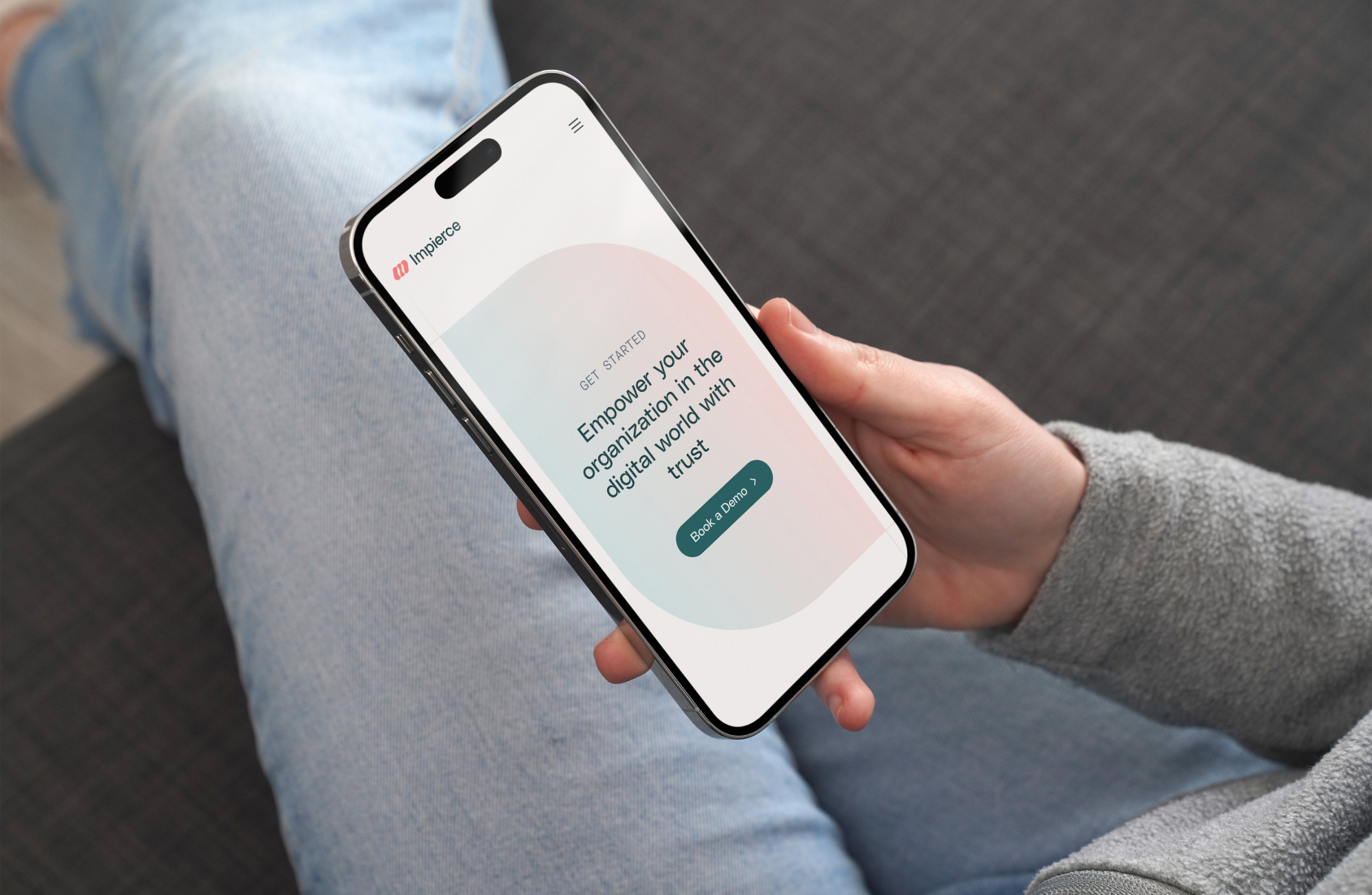 A person holding a smartphone with a screen displaying a website that says, "Empower your organization in the digital world with trust" and a "Get in contact" button. The person is seated on a gray couch, wearing light blue jeans.