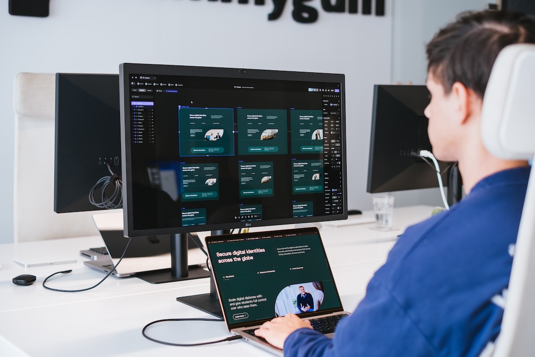 A person sits at a desk using a laptop connected to a large monitor displaying dark-themed dashboard screens with various data panels and graphics in a modern office setting.