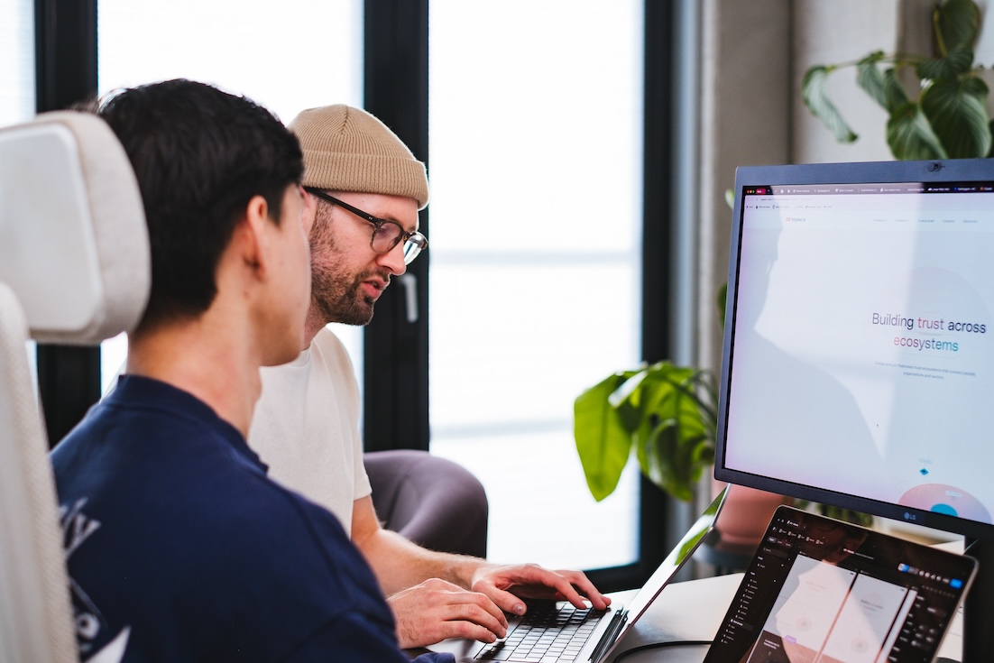 Two people sit at a desk working together on laptops. One person wears a beige beanie and glasses, while the other has dark hair. A large monitor displays a website. A plant and large windows are in the background.