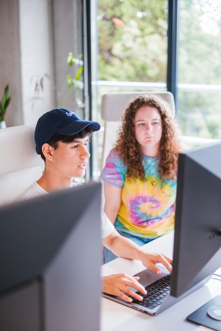 Two people sit side by side at a desk with computer monitors. One, wearing a dark cap, is typing on a laptop, while the other, in a colorful tie-dye shirt, looks at the screen. A window and greenery are visible behind them.