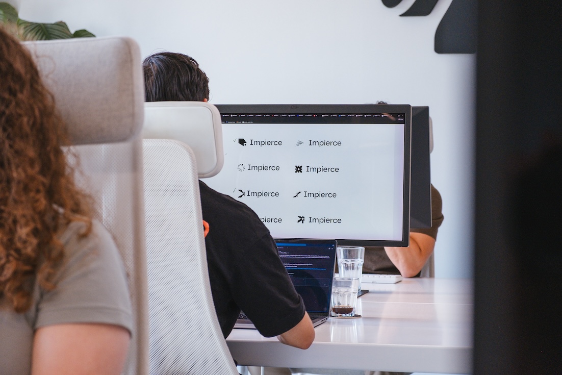 Three people sit at desks in an office, partially obscured by computer monitors. One person is typing, while another has a glass of water and a laptop in front of them. A website or app is displayed on the main monitor.