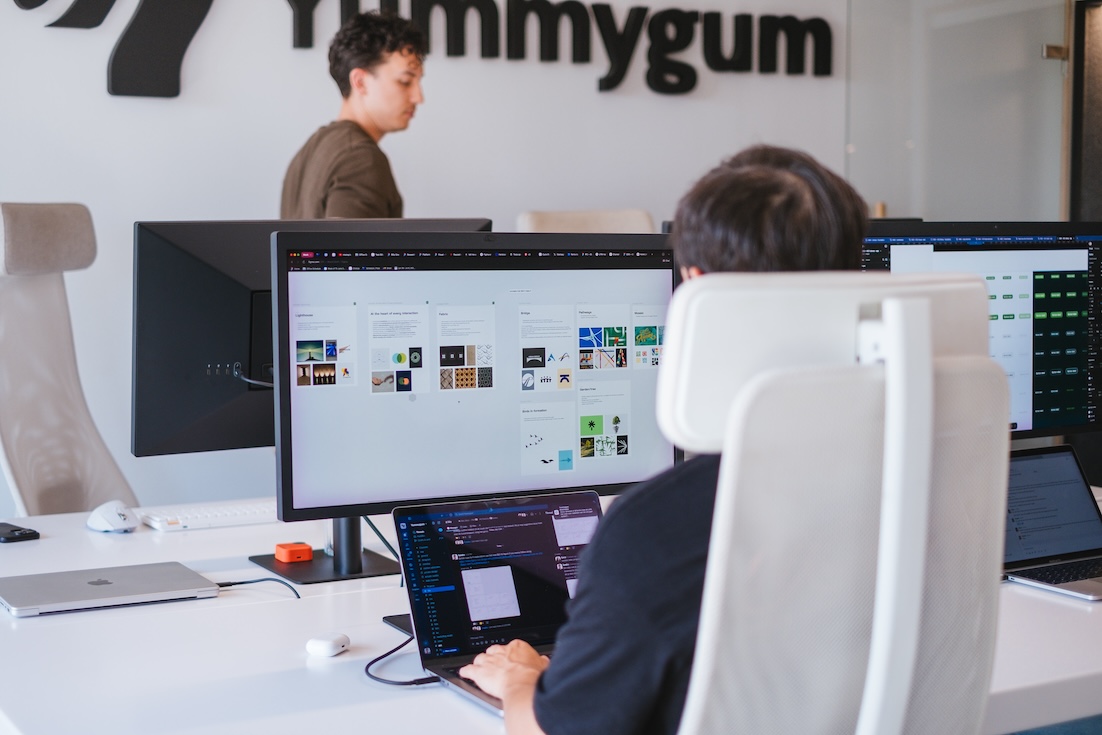 Two people work in a modern office; one types on a keyboard at a large monitor displaying digital design projects, while another stands in the background near a wall sign that reads “Yummygum.”.